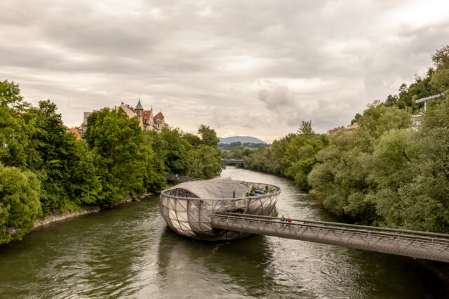 Blick auf die Murinsel in Graz an einem bewölkten Sommertag – modernes, schalenförmiges Bauwerk auf der Mur, umgeben von grünen Bäumen, historischer Architektur im Hintergrund und Spaziergänger*innen auf der Brücke