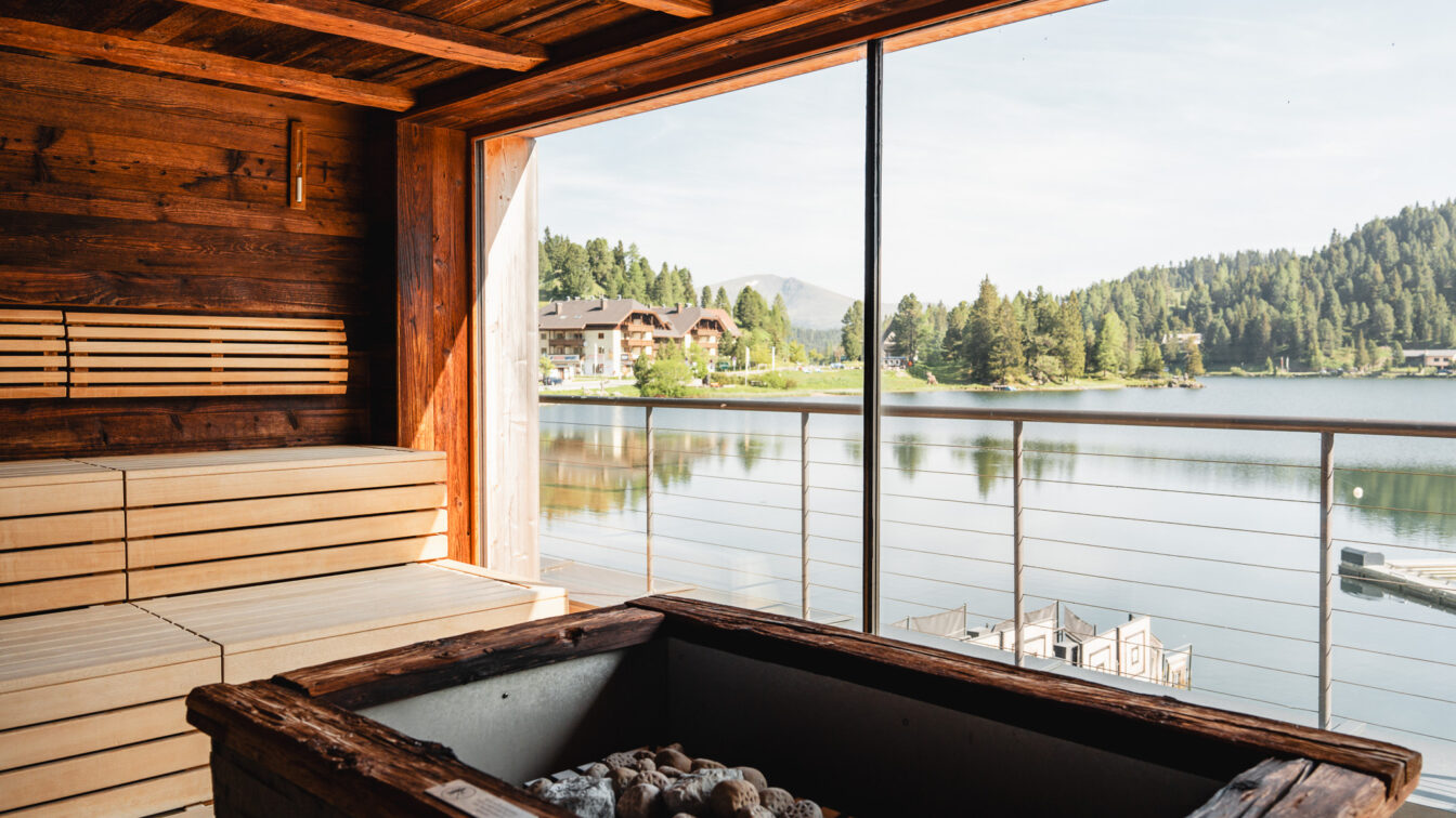 Blick aus einer Sauna heraus mit Glas-Wand und Sicht auf einen See mit Bergen im Hintergrund.