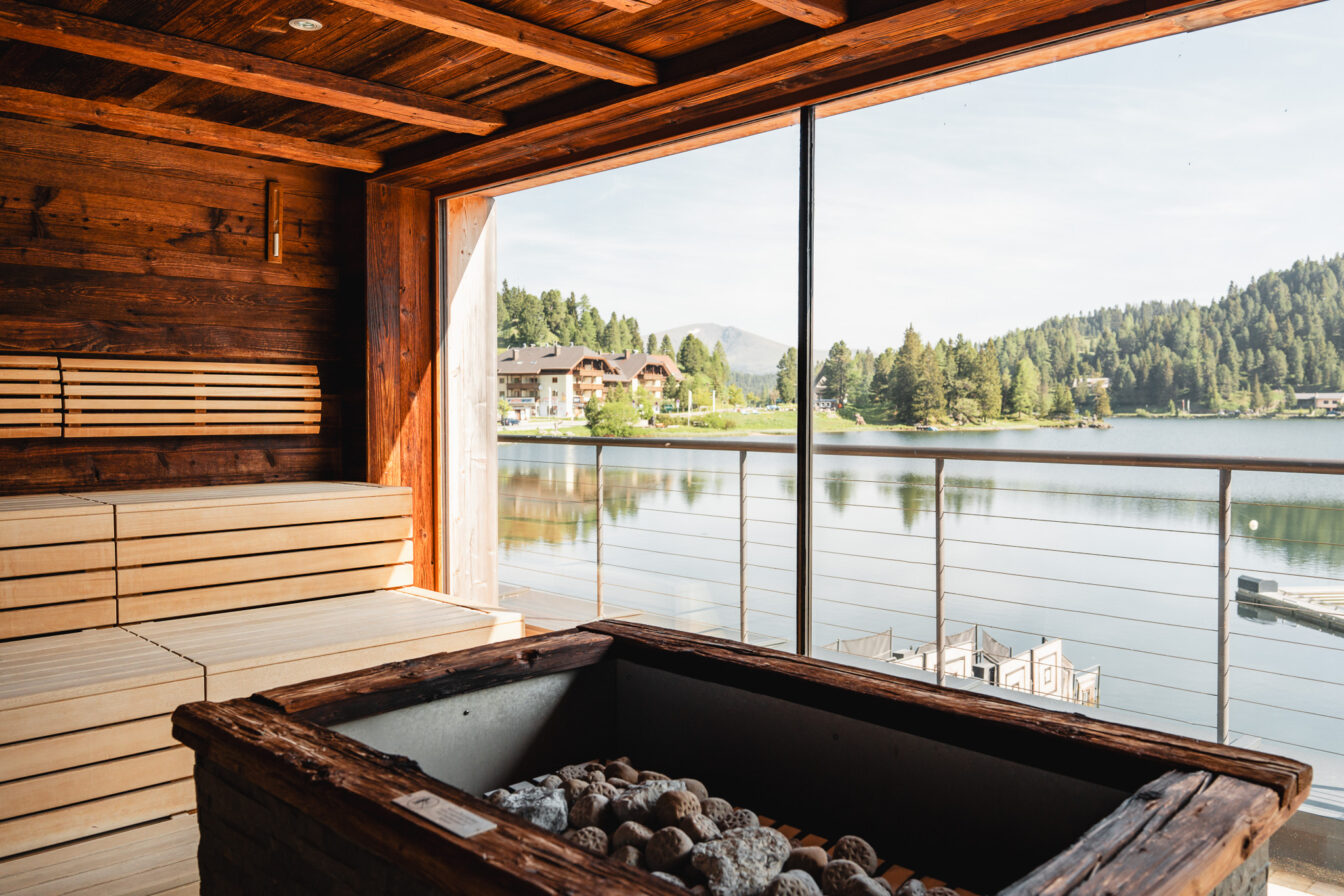 Blick aus einer Sauna heraus mit Glas-Wand und Sicht auf einen See mit Bergen im Hintergrund.