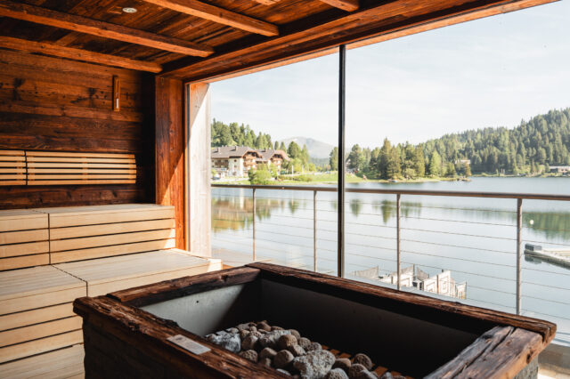 Blick aus einer Sauna heraus mit Glas-Wand und Sicht auf einen See mit Bergen im Hintergrund.