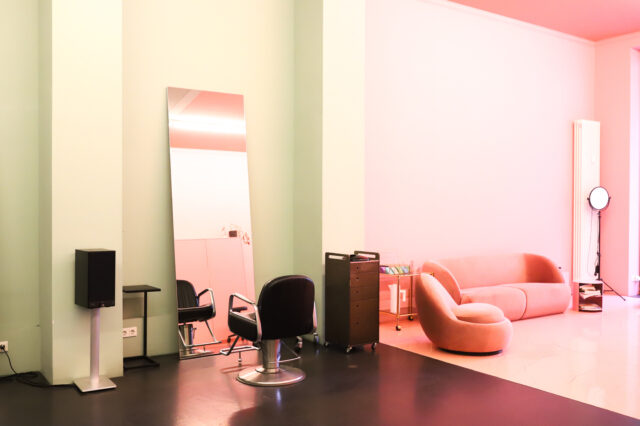 Foto eines Friseur in Berlin mit minimalistischer Einrichtung. Man sieht einen Friseurstuhl vor einem an der Wand gelehnten Spiegel und eine Couch daneben.