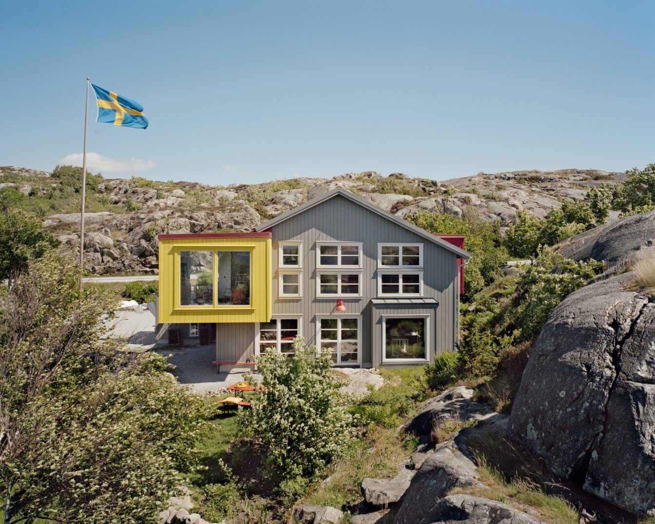 Modernes Gästehaus Lådfabriken an der schwedischen Westküste – stilvolles Architektenhaus mit farbigen Akzenten inmitten felsiger Landschaft und Natur, mit schwedischer Flagge im Vordergrund.