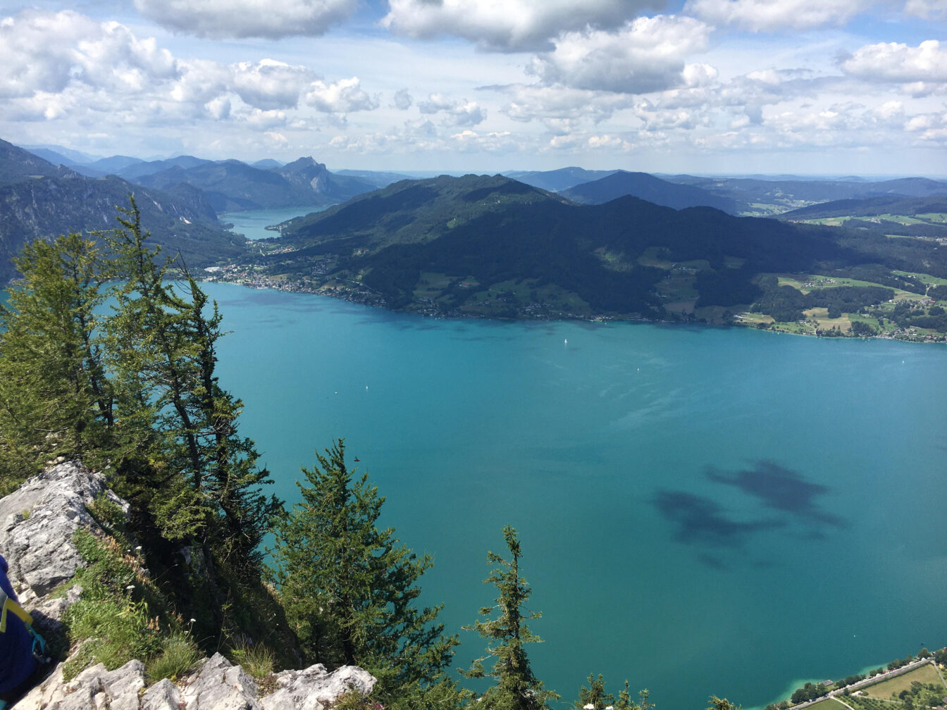 Aussichtsreicher Blick vom Gipfel des Mahdlgupf über den türkisfarbenen Attersee in Oberösterreich. Im Vordergrund felsiger Abgrund mit Latschen und Nadelbäumen. Im Hintergrund eine weite Berglandschaft mit bewaldeten Hügeln, kleinen Ortschaften am Seeufer und einem wolkigen Himmel, der sich im Wasser spiegelt.