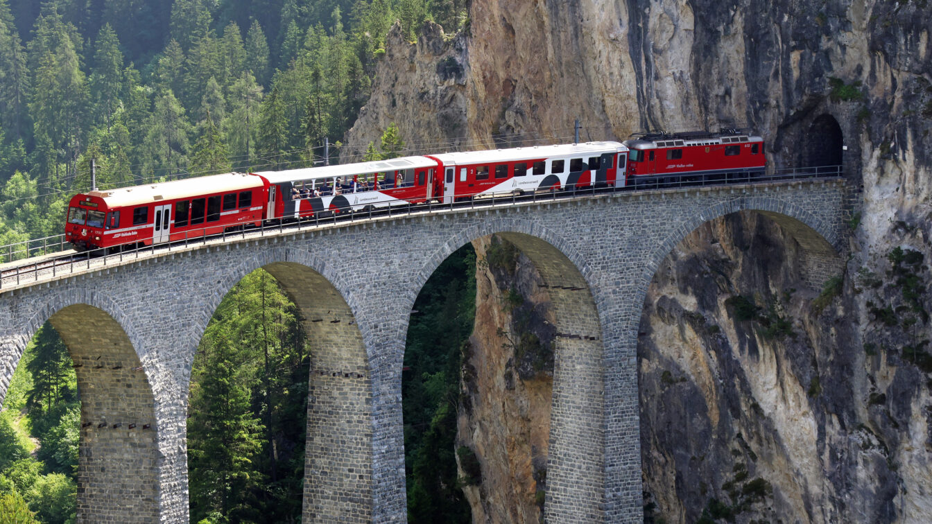 Ein rot-weißer Zug, der vorbei an einer grünen Landschaft über ein Landwasserviadukt fährt.