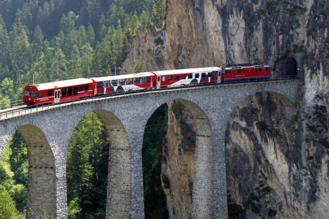 Ein rot-weißer Zug, der vorbei an einer grünen Landschaft über ein Landwasserviadukt fährt.
