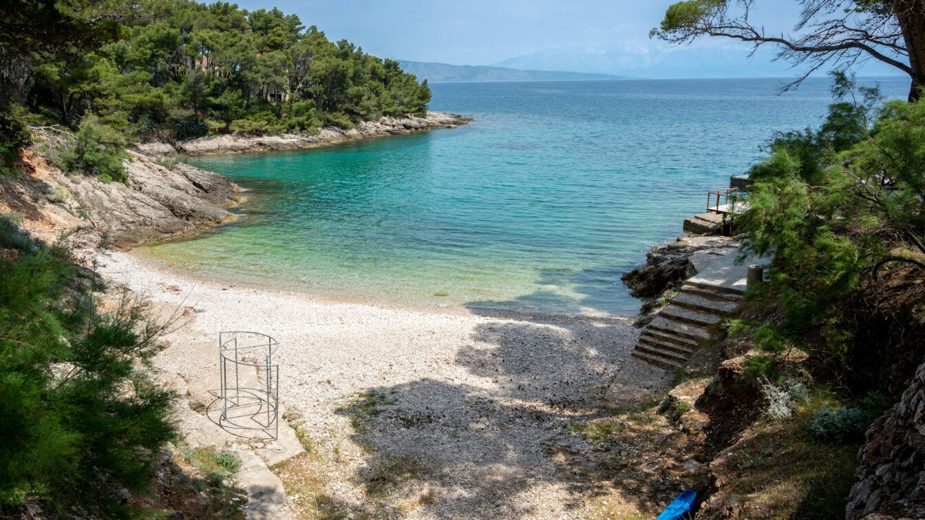 Ein menschenleerer Strand in Kroatien, der umringt von Bäumen türkisblaues Wasser verspricht.