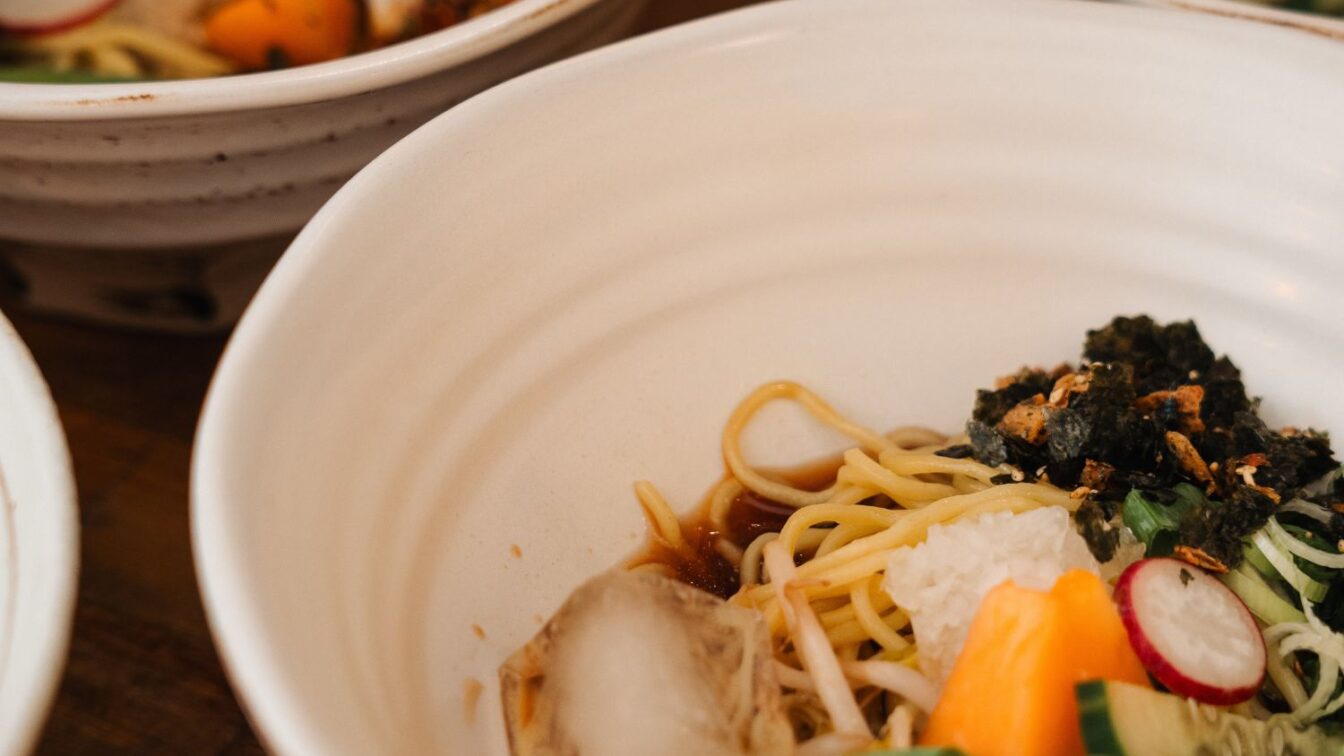Drei weiße Keramikschalen mit modernen Ramen-Gerichten: feine Weizennudeln in aromatischer Brühe, garniert mit knusprigem Seetang, Radieschenscheiben, Frühlingszwiebeln und buntem Gemüse – zeitgemäße japanische Küche in stilvollem Ambiente.