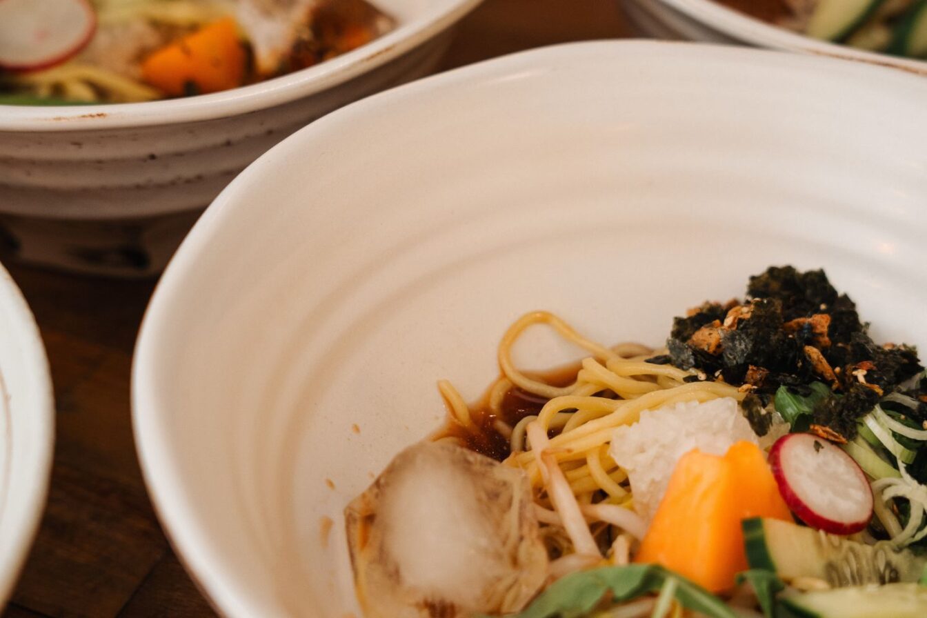 Drei weiße Keramikschalen mit modernen Ramen-Gerichten: feine Weizennudeln in aromatischer Brühe, garniert mit knusprigem Seetang, Radieschenscheiben, Frühlingszwiebeln und buntem Gemüse – zeitgemäße japanische Küche in stilvollem Ambiente.