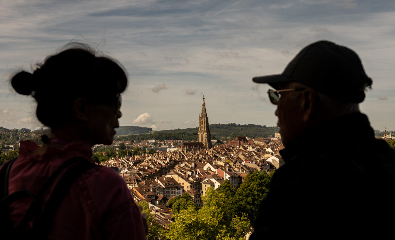 Zwei Personen im Schatten betrachten den Panoramablick über die Altstadt von Bern mit der markanten Berner Münsterkirche im Zentrum, aufgenommen vom Aussichtspunkt beim Restaurant Rosengarten.