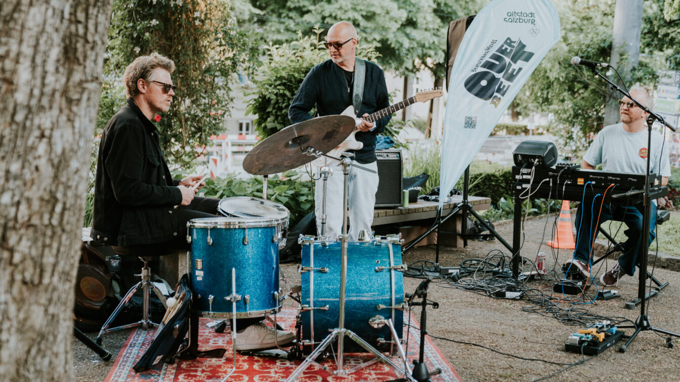Live-Musik im Schanzlpark Salzburg – Band mit Schlagzeug, Gitarre und Keyboard bei Open-Air-Konzert der Veranstaltungsreihe 'Altstadt Salzburg Über d’Gass', stimmungsvolle Atmosphäre unter freiem Himmel.