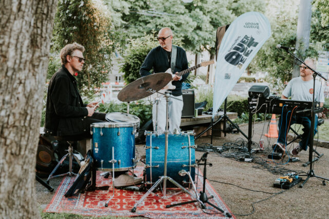 Live-Musik im Schanzlpark Salzburg – Band mit Schlagzeug, Gitarre und Keyboard bei Open-Air-Konzert der Veranstaltungsreihe 'Altstadt Salzburg Über d’Gass', stimmungsvolle Atmosphäre unter freiem Himmel.