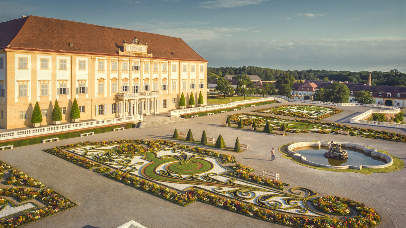 Blick auf das barocke Schloss Hof mit seiner prachtvollen Fassade und dem kunstvoll angelegten Schlossgarten. Der symmetrische Garten ist mit blühenden Blumenbeeten, Hecken, Skulpturen und einem Springbrunnen gestaltet. Die Szene ist in warmes Sonnenlicht getaucht und vermittelt eine Atmosphäre historischer Eleganz und Ruhe.