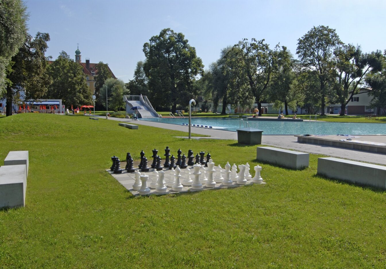 Ein Schachbrett mit großen Spielfiguren auf einer grünen Wiese mit Freibad in München im Hintergrund.