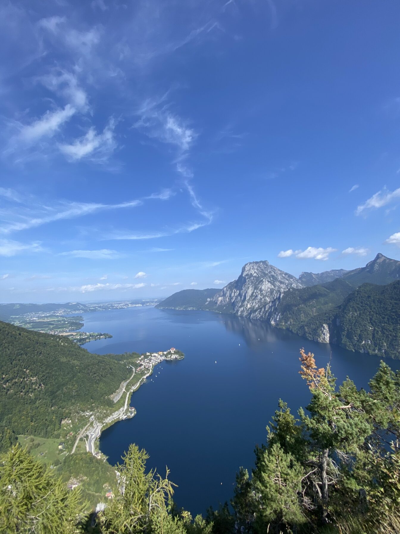 Panoramablick vom Kleinen oder Großen Sonnstein über den tiefblauen Traunsee in Oberösterreich. Im Vordergrund grüne Baumwipfel, darunter schlängelt sich eine Straße entlang des Seeufers. In der Bildmitte die Halbinsel mit der Ortschaft Traunkirchen. Im Hintergrund ragt der mächtige Traunstein markant empor, eingerahmt von weiteren bewaldeten Bergzügen unter einem strahlend blauen Himmel mit zarten Schleierwolken.