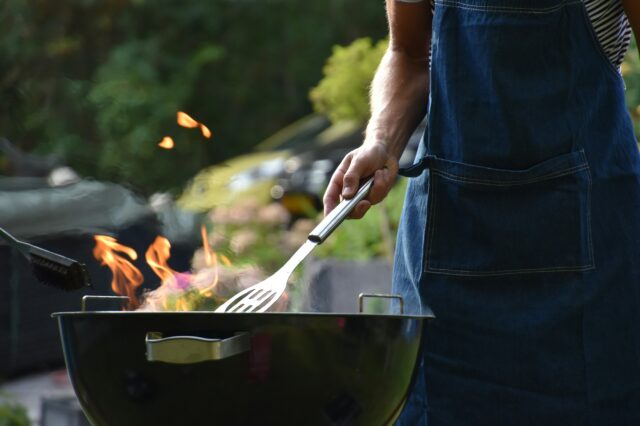 Eine Person mit Grillschürze und Wender steht an einem Feuer-Griller