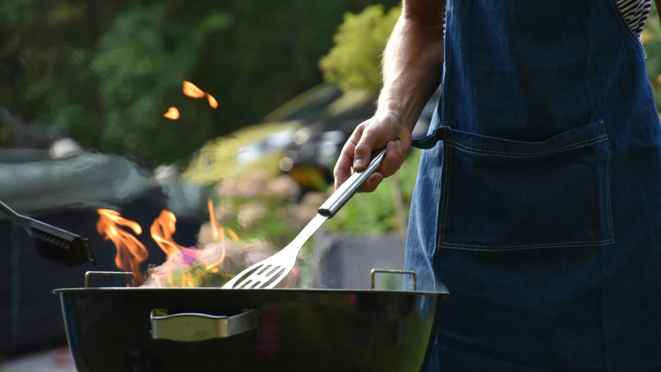 Eine Person mit Grillschürze und Wender steht an einem Feuer-Griller