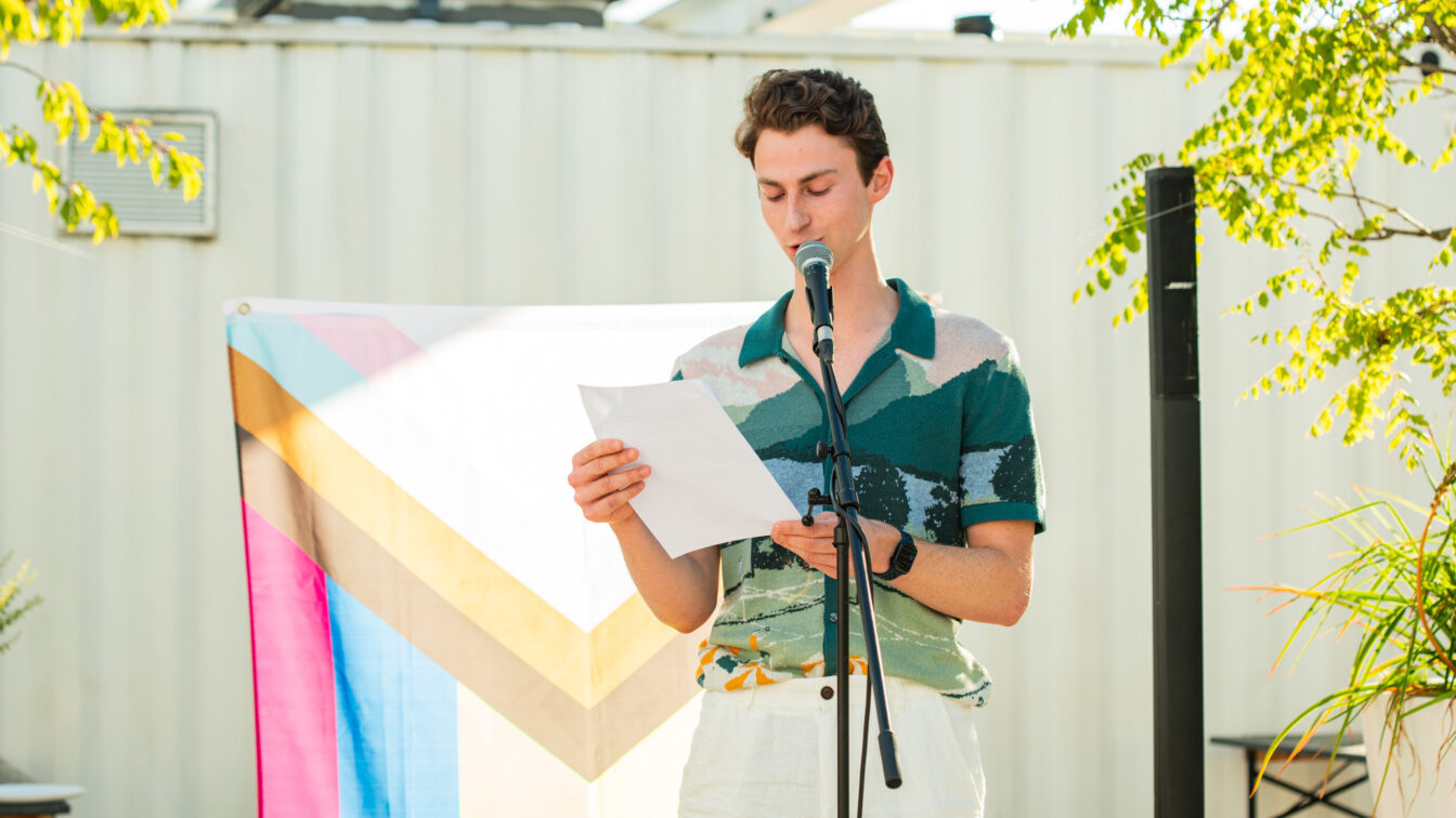 Person mit buntem Hemd auf einer flachen Bühne, die einen Text liest, mit Pride-Flagge im Hintergrund.
