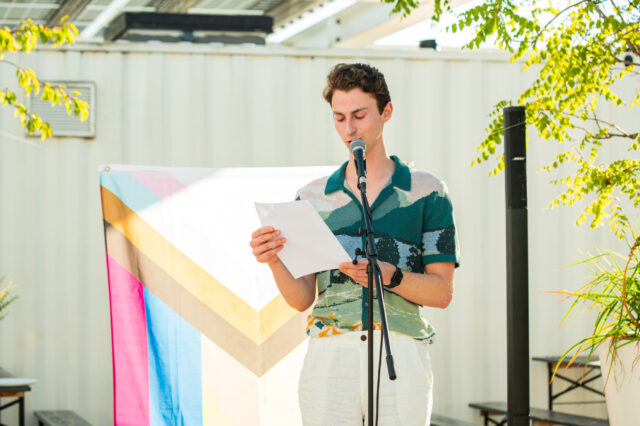 Person mit buntem Hemd auf einer flachen Bühne, die einen Text liest, mit Pride-Flagge im Hintergrund.