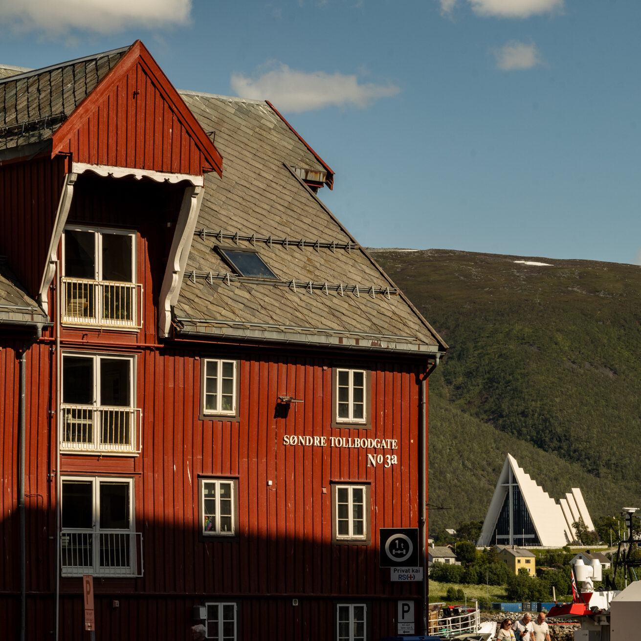 Historisches, rot gestrichenes Holzgebäude mit grauem Schieferdach in Tromsø, markiert mit der Adresse "Søndre Tollbodgate No 3a", dem Sitz des Polarmuseums. Im Hintergrund ist die moderne Eismeerkathedrale zwischen grün bewaldeten Hängen zu sehen. Einige Menschen gehen bei sonnigem Wetter an der Hafenpromenade entlang, darüber ein klarer Himmel mit wenigen Wolken.