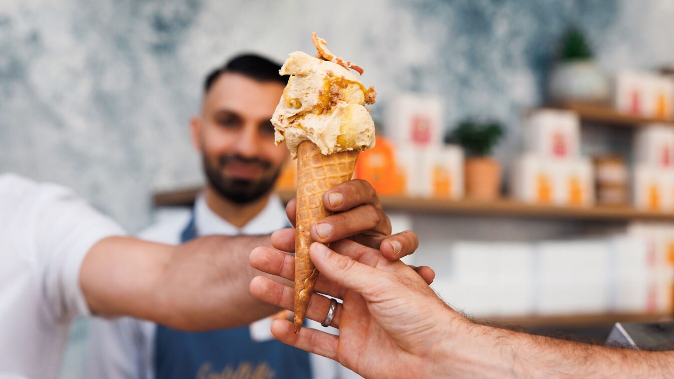 Eine Hand, die eine Kugel Eis in der Waffel überreicht mit einem Mann im Hintergrund.