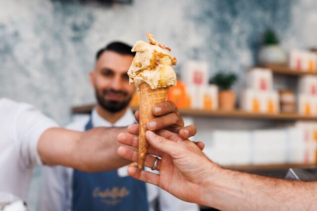 Eine Hand, die eine Kugel Eis in der Waffel überreicht mit einem Mann im Hintergrund.