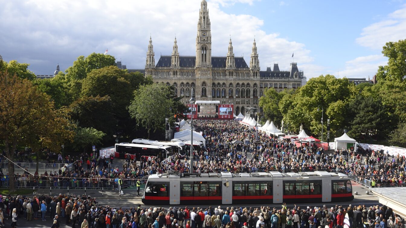Große Menschenmenge bei der Tram-WM vor dem Wiener Rathaus, zahlreiche Besucher versammeln sich rund um eine moderne Straßenbahn und mehrere Infostände, im Hintergrund das historische Rathausgebäude mit seinen Türmen