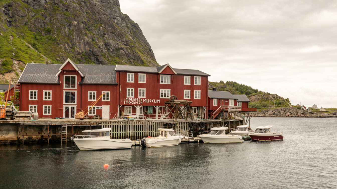 Å i Lofoten, Norwegen – historische rote Holzhäuser am Wasser mit dem Norwegischen Stockfischmuseum, umgeben von steilen Felswänden und Booten im ruhigen Hafen bei bewölktem Himmel.