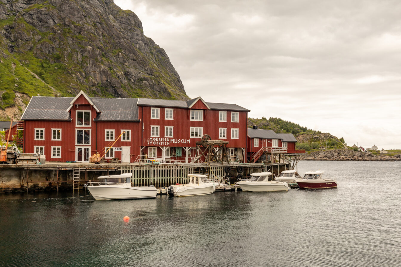 Å i Lofoten, Norwegen – historische rote Holzhäuser am Wasser mit dem Norwegischen Stockfischmuseum, umgeben von steilen Felswänden und Booten im ruhigen Hafen bei bewölktem Himmel.