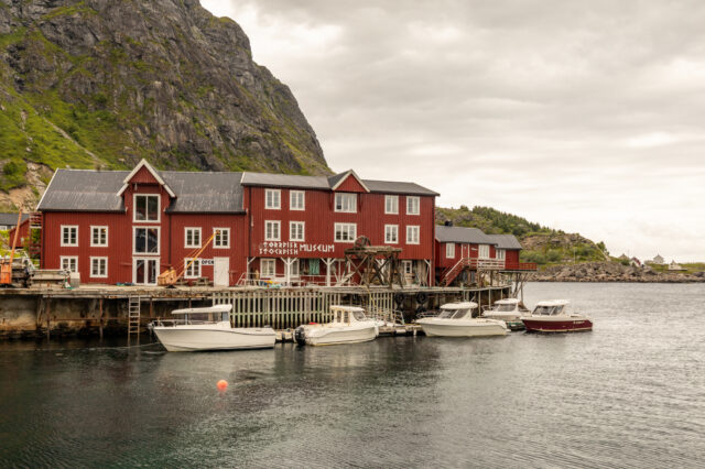 Å i Lofoten, Norwegen – historische rote Holzhäuser am Wasser mit dem Norwegischen Stockfischmuseum, umgeben von steilen Felswänden und Booten im ruhigen Hafen bei bewölktem Himmel.