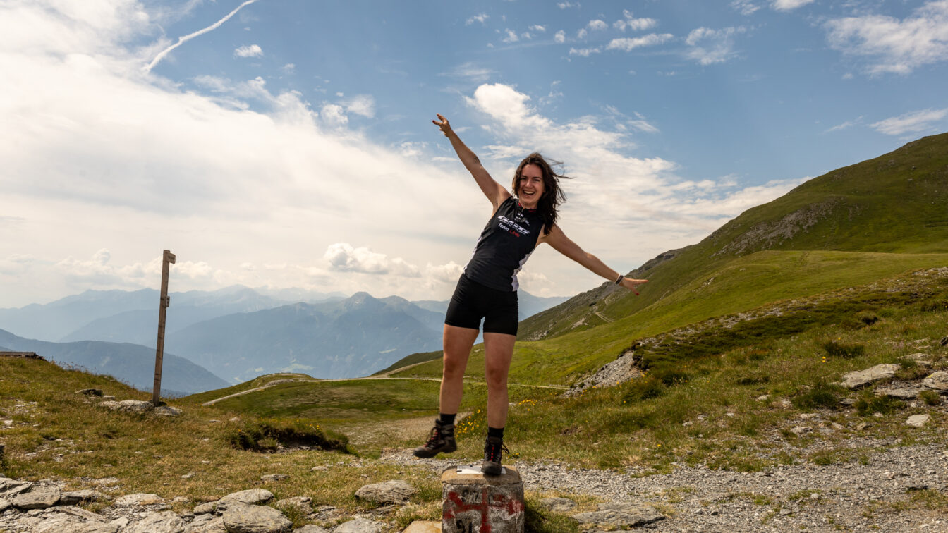 Person in Sportkleidung steht lachend auf einem Grenzstein am Pass während der Alpenüberquerung, umgeben von grünen Hügeln und einem weiten Bergpanorama unter blauem Himmel mit leichten Wolken.