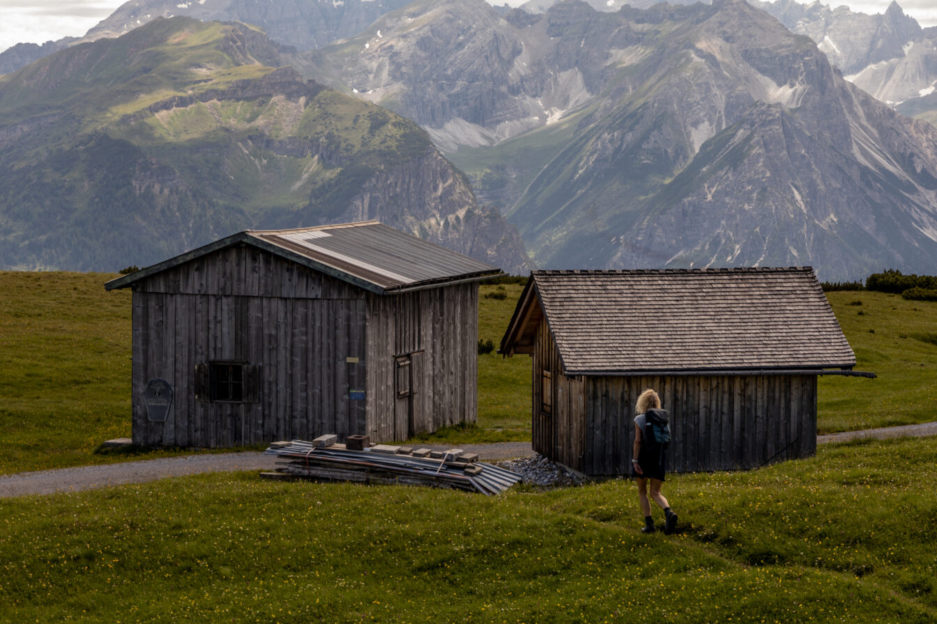 Person mit Rucksack wandert auf einer Almwiese an zwei kleinen, einfachen Holzhütten vorbei, im Hintergrund ragen steile, grüne und felsige Alpen während einer Alpenüberquerung in die Höhe.