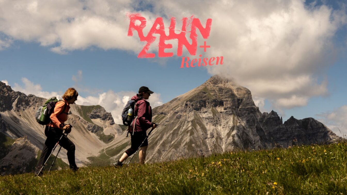 Zwei wandernde Personen mit Rucksäcken und Wanderstöcken überqueren eine grüne Wiese vor einer beeindruckenden Bergkulisse mit steilen Felswänden und Wolken am Himmel. Im oberen Bildbereich steht in roter Schrift „Rauzen + Reisen“.