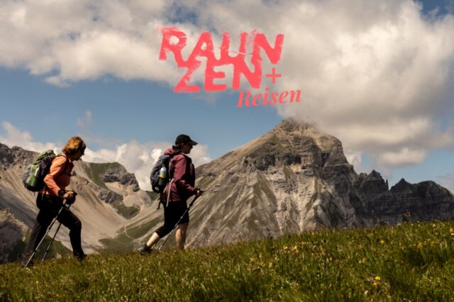 Zwei wandernde Personen mit Rucksäcken und Wanderstöcken überqueren eine grüne Wiese vor einer beeindruckenden Bergkulisse mit steilen Felswänden und Wolken am Himmel. Im oberen Bildbereich steht in roter Schrift „Rauzen + Reisen“.
