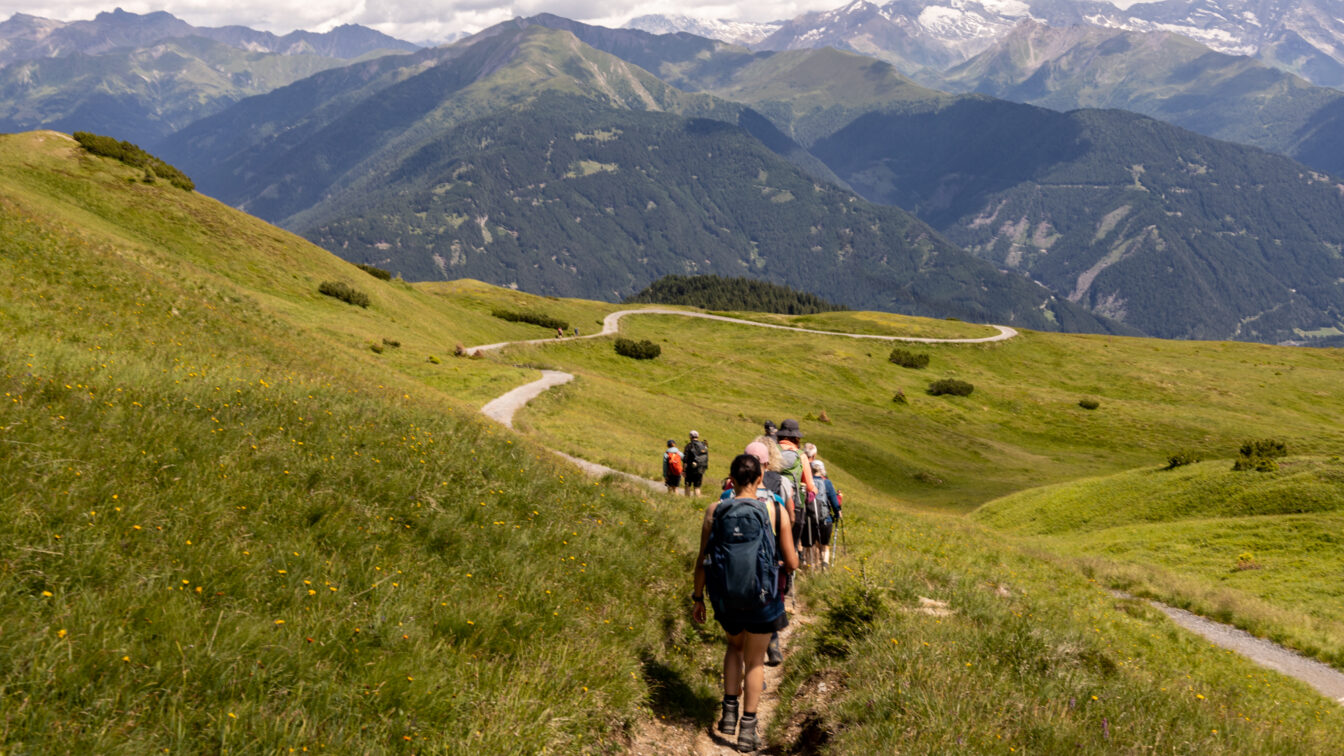 Wandergruppe mit Rucksäcken auf schmalem Bergpfad bei der Alpenüberquerung von Garmisch nach Sterzing, grünes Almwiesenpanorama mit sanften Hügeln, dahinter hohe Berge mit Schneefeldern unter bewölktem Himmel.