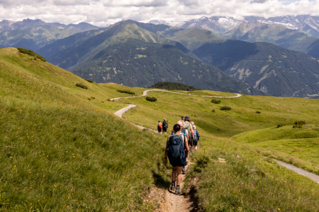 Wandergruppe mit Rucksäcken auf schmalem Bergpfad bei der Alpenüberquerung von Garmisch nach Sterzing, grünes Almwiesenpanorama mit sanften Hügeln, dahinter hohe Berge mit Schneefeldern unter bewölktem Himmel.