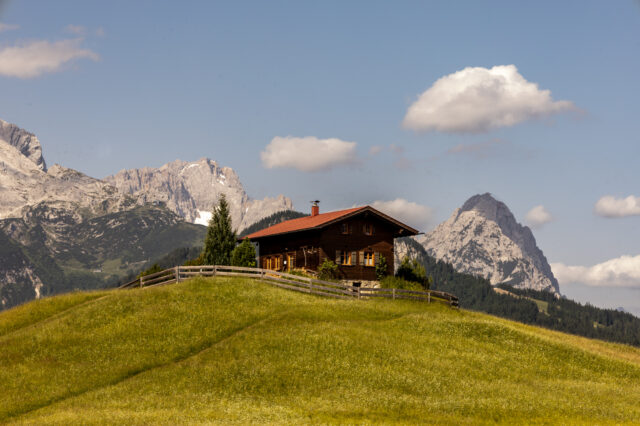 Holzhaus auf einer grünen Almwiese bei Garmisch-Partenkirchen, im Hintergrund schroffe Alpen mit einzelnen Schneefeldern und blauer Himmel mit Wolken, idealer Startpunkt für eine Alpenüberquerung nach Mittenwald.