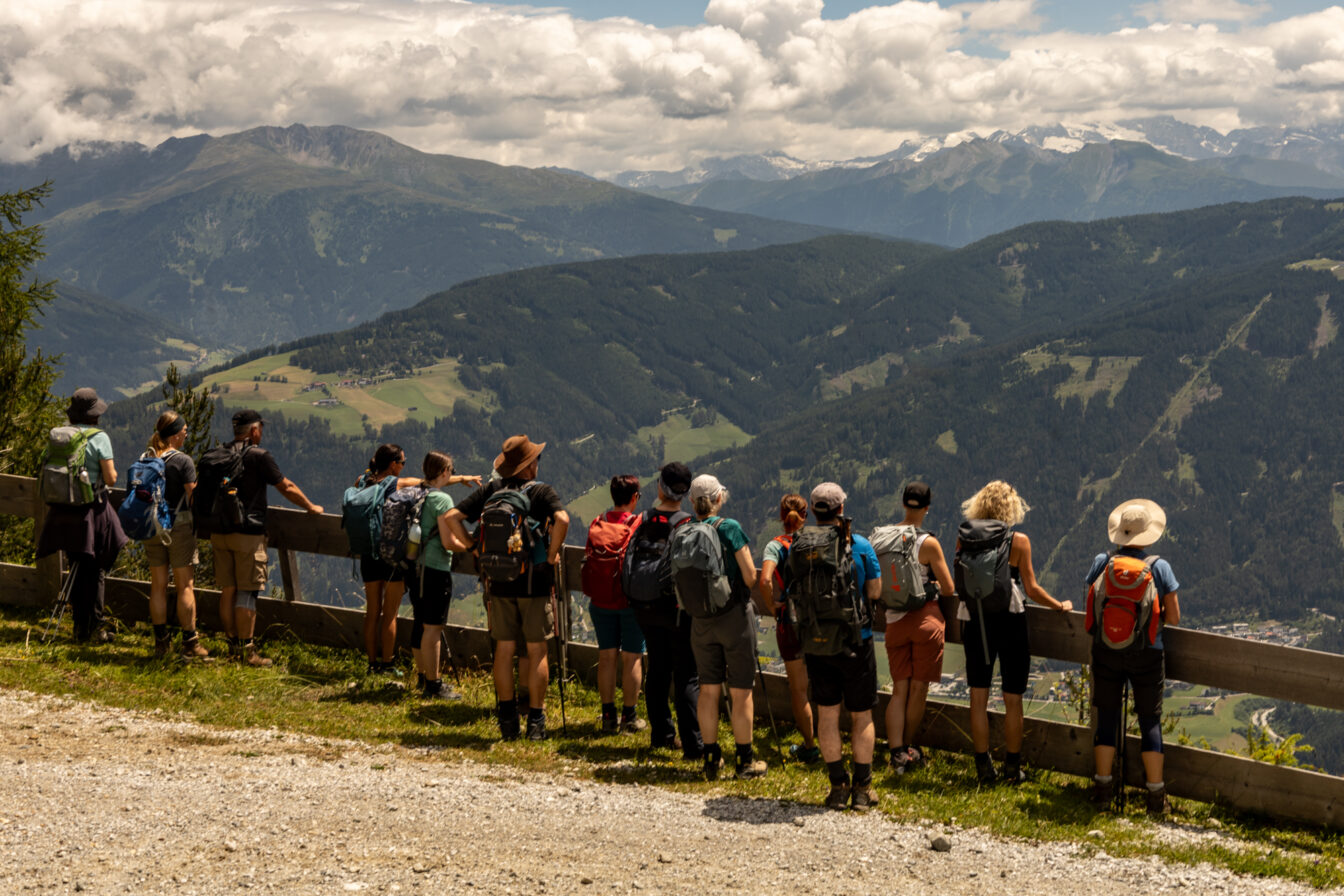 Eine Gruppe von Wandernden mit Rucksäcken steht an einem Holzgeländer und blickt ins grüne Tal und auf Berge mit Wolken und Schneefeldern im Hintergrund.