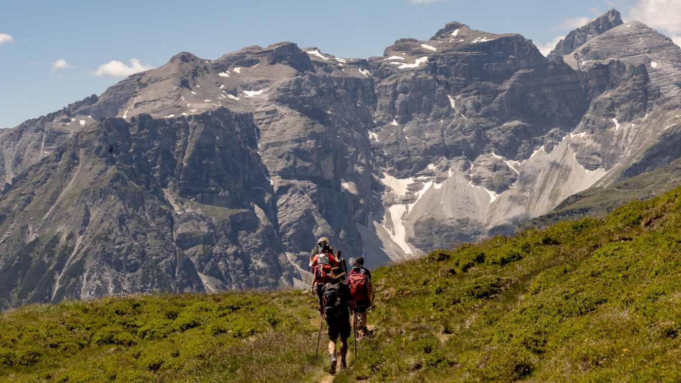 Wandergruppe mit Rucksäcken und Stöcken auf einem schmalen Almweg, der auf ein mächtiges, graues Felsmassiv mit Schneefeldern und blauem Himmel in den Tiroler Alpen zuführt.
