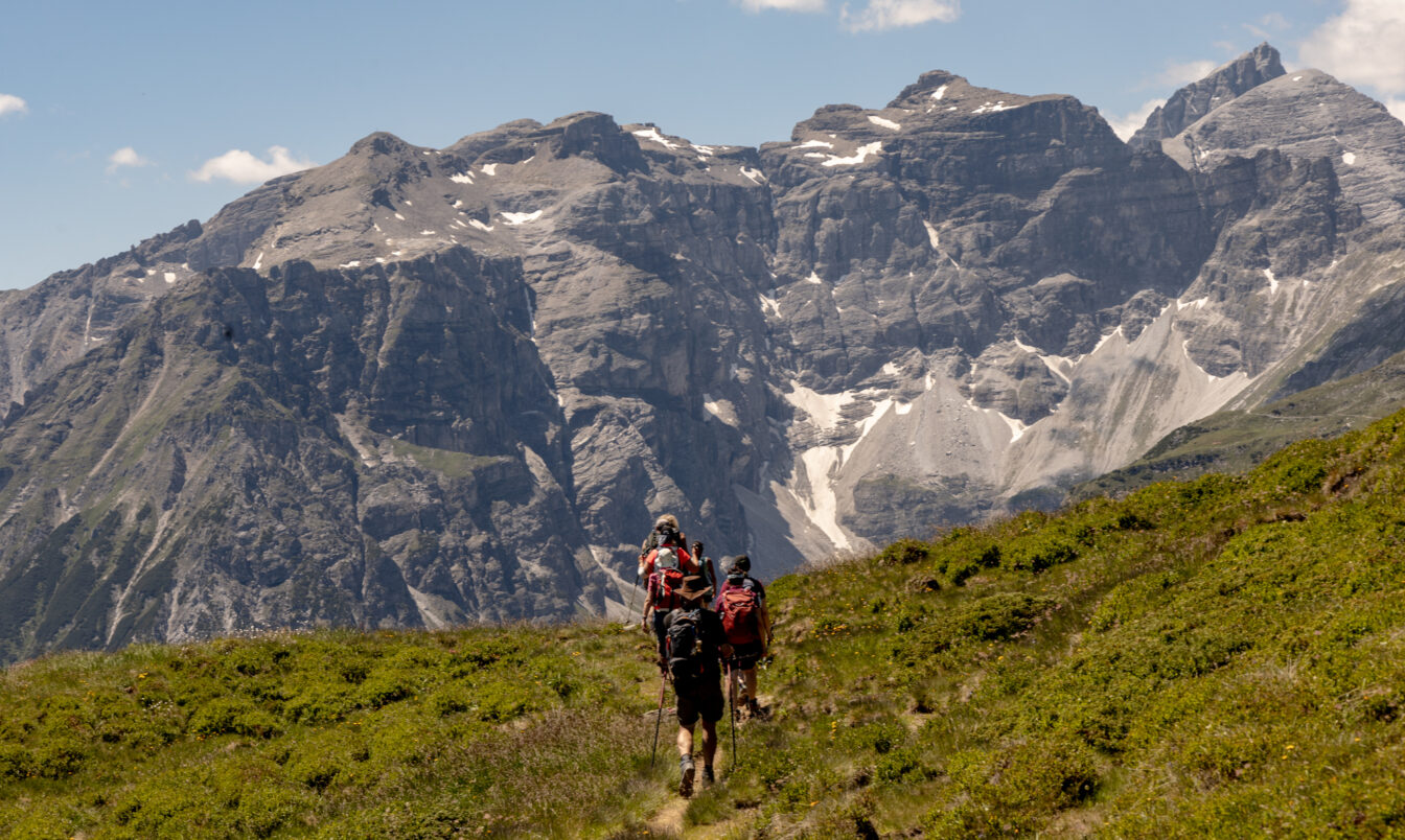 Wandergruppe mit Rucksäcken und Stöcken auf einem schmalen Almweg, der auf ein mächtiges, graues Felsmassiv mit Schneefeldern und blauem Himmel in den Tiroler Alpen zuführt.
