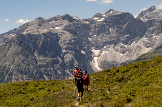 Wandergruppe mit Rucksäcken und Stöcken auf einem schmalen Almweg, der auf ein mächtiges, graues Felsmassiv mit Schneefeldern und blauem Himmel in den Tiroler Alpen zuführt.