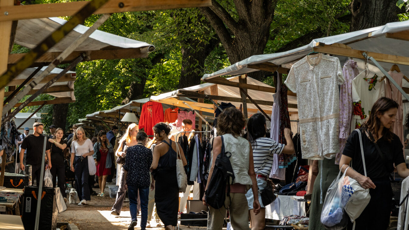 Belebter Second-Hand-Markt in Berlin unter großen schattenspendenden Bäumen. Zahlreiche Menschen schlendern entlang der Stände mit Kleidung, die an Holzgestellen hängt. Die Atmosphäre wirkt entspannt und sommerlich, mit vielfältiger Mode und neugierigen Besucher*innen.