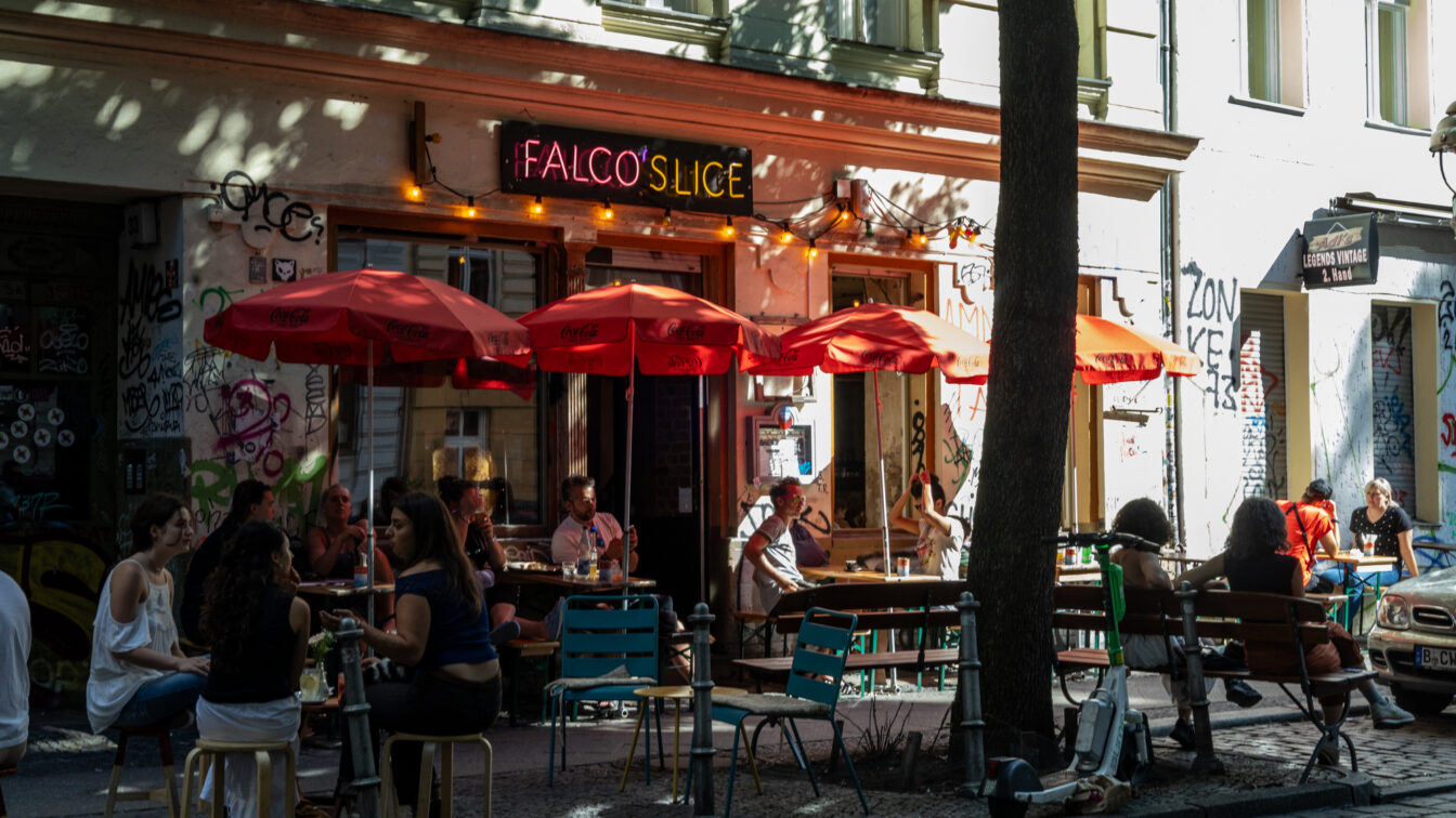 Sommerszene vor der Pizzeria ‚Falco’s Slice‘ in Berlin: Menschen sitzen entspannt unter roten Sonnenschirmen auf dem Gehweg, umgeben von Graffiti, bunten Stühlen und urbanem Flair – ein typischer Moment für einen Berlin Urlaub abseits der Touristenpfade.