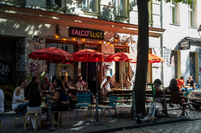 Sommerszene vor der Pizzeria ‚Falco’s Slice‘ in Berlin: Menschen sitzen entspannt unter roten Sonnenschirmen auf dem Gehweg, umgeben von Graffiti, bunten Stühlen und urbanem Flair – ein typischer Moment für einen Berlin Urlaub abseits der Touristenpfade.