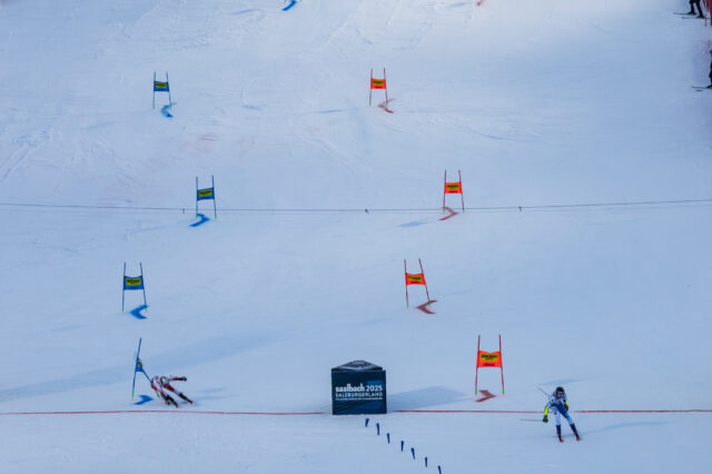 Zwei alpine Skirennläufer:innen durchfahren parallel einen Riesenslalom-Parcours während eines Qualifikationsrennens oder Wettbewerbs. Im Zielbereich steht ein Schild mit der Aufschrift „Saalbach 2025 SalzburgerLand“, dem Austragungsort der FIS Alpinen Ski-Weltmeisterschaften 2025, die auch als Vorbereitung auf die Olympischen Winterspiele 2026 in Mailand-Cortina dienen.