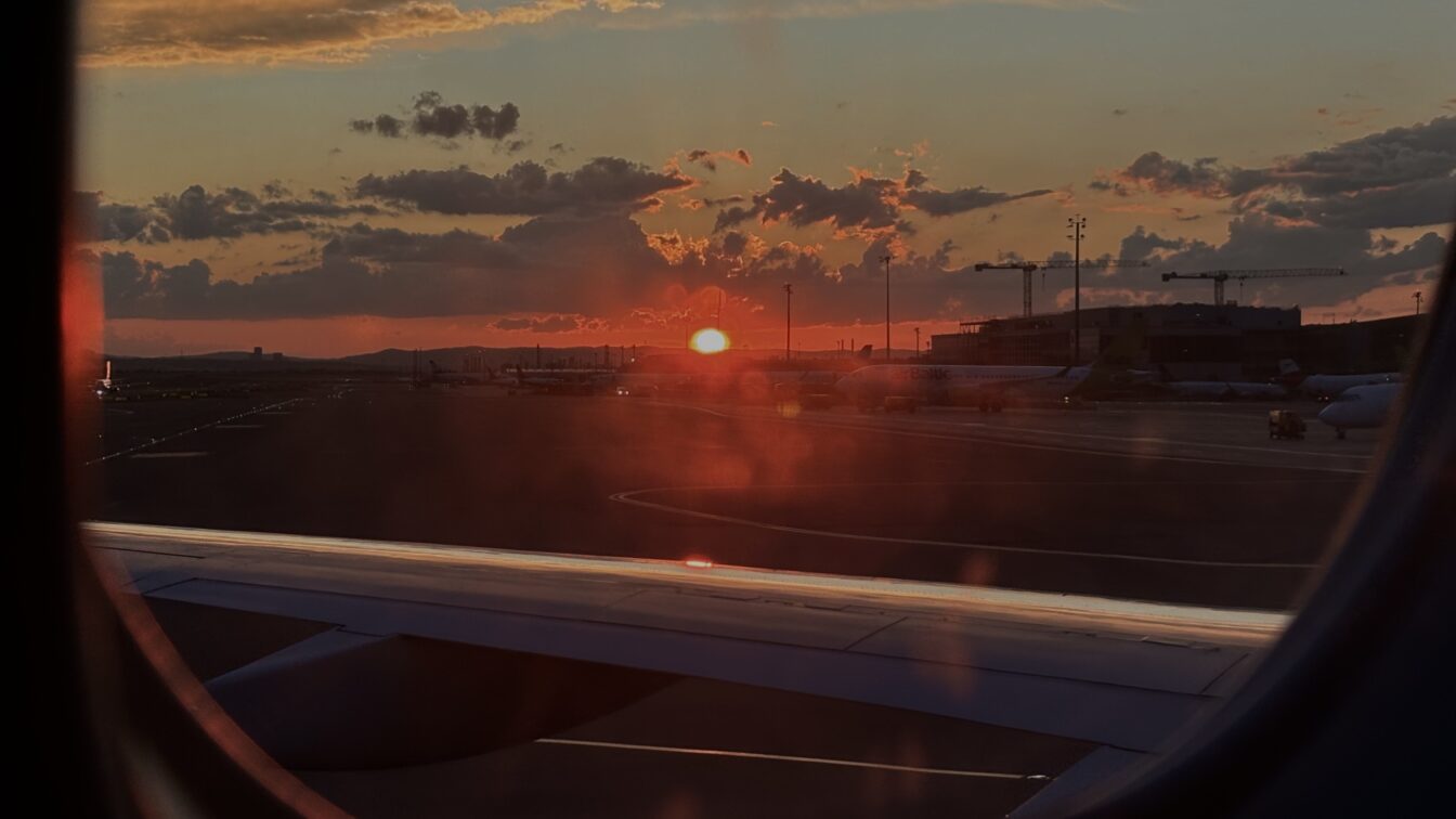 Blick aus dem Fenster eines Flugzeugs beim Sonnenuntergang auf dem Rollfeld eines Flughafens. Die Szene zeigt parkende Flugzeuge, den Flughafenbetrieb mit Gebäuden und Kränen im Hintergrund. Das Bild symbolisiert eine Wartezeit oder mögliche Flugverspätung – ein typischer Anlass, bei dem Passagieren Fluggastrechte zustehen können, etwa Entschädigungen bei verspäteten oder gestrichenen Flügen.