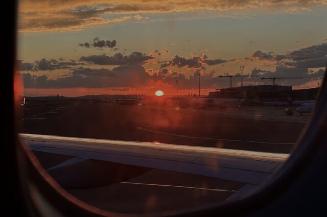 Blick aus dem Fenster eines Flugzeugs beim Sonnenuntergang auf dem Rollfeld eines Flughafens. Die Szene zeigt parkende Flugzeuge, den Flughafenbetrieb mit Gebäuden und Kränen im Hintergrund. Das Bild symbolisiert eine Wartezeit oder mögliche Flugverspätung – ein typischer Anlass, bei dem Passagieren Fluggastrechte zustehen können, etwa Entschädigungen bei verspäteten oder gestrichenen Flügen.