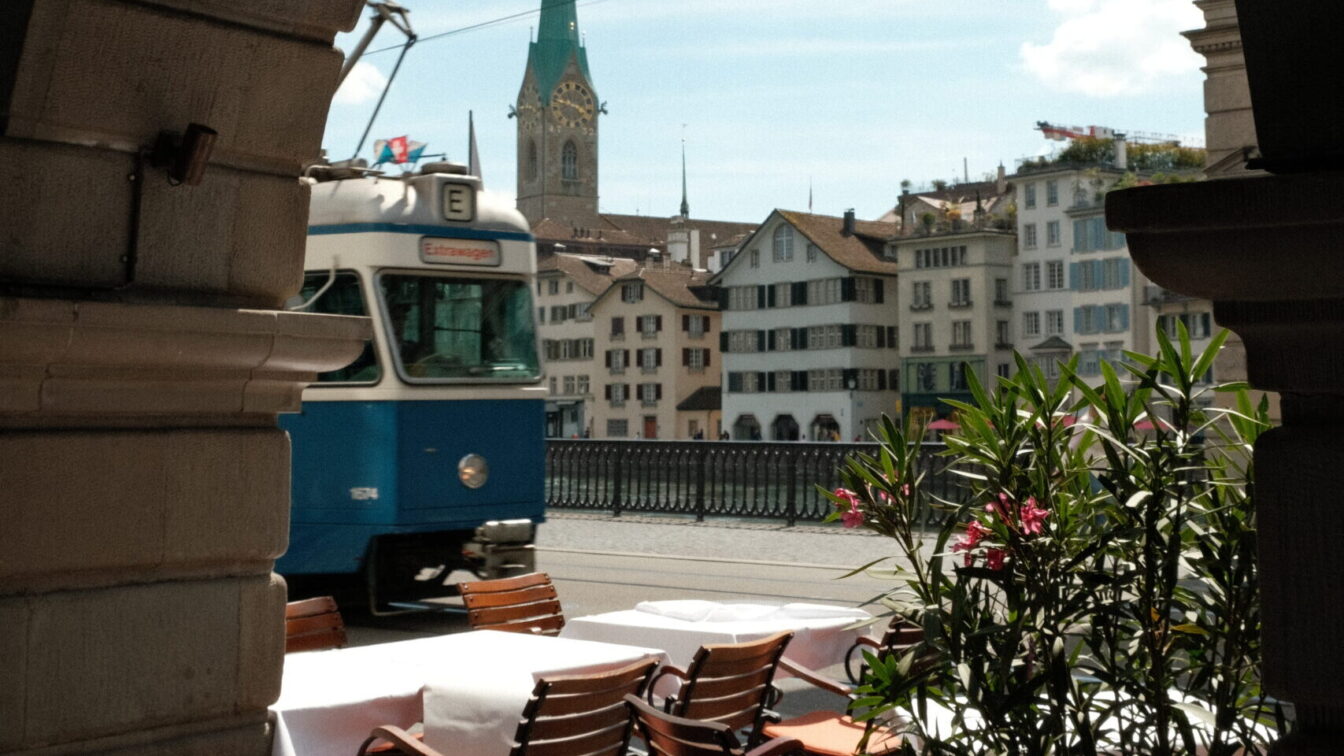 Blick durch einen steinernen Torbogen in Zürich auf einen blauen Tram, dahinter die Fraumünsterkirche mit ihrem markanten grünen Turm. Im Vordergrund stehen gedeckte Restauranttische mit Stühlen auf einer Terrasse am Fluss.