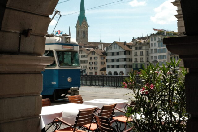 Blick durch einen steinernen Torbogen in Zürich auf einen blauen Tram, dahinter die Fraumünsterkirche mit ihrem markanten grünen Turm. Im Vordergrund stehen gedeckte Restauranttische mit Stühlen auf einer Terrasse am Fluss.