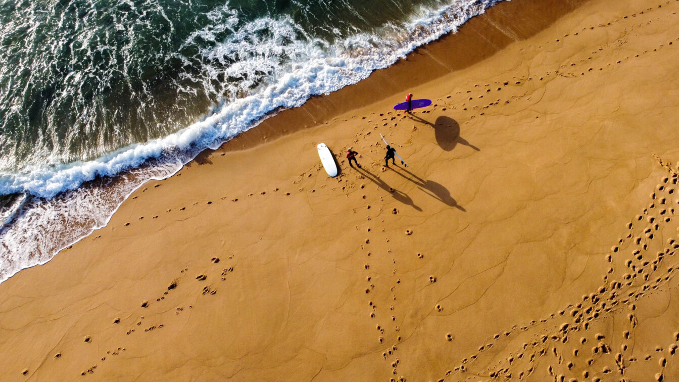 Vogelperspektive auf drei surfende Personen am Strand vor dem Wasser in Portugal
