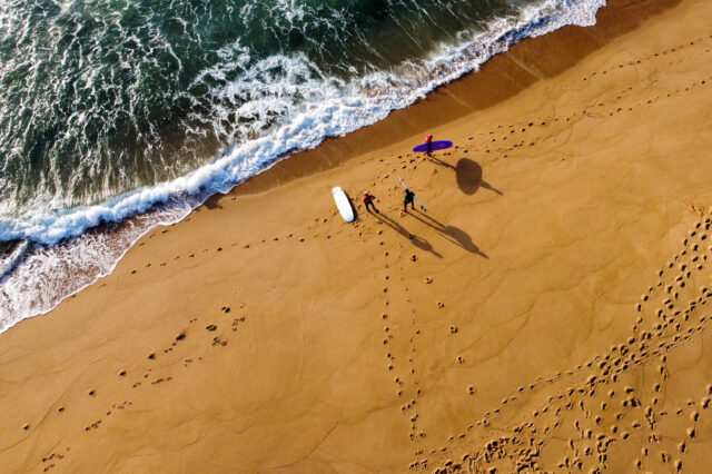 Vogelperspektive auf drei surfende Personen am Strand vor dem Wasser in Portugal